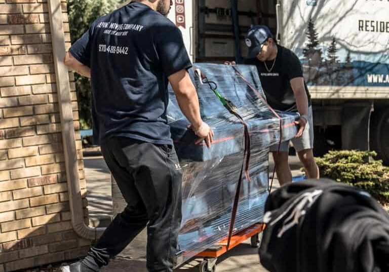 Movers transporting wrapped furniture with a dolly.